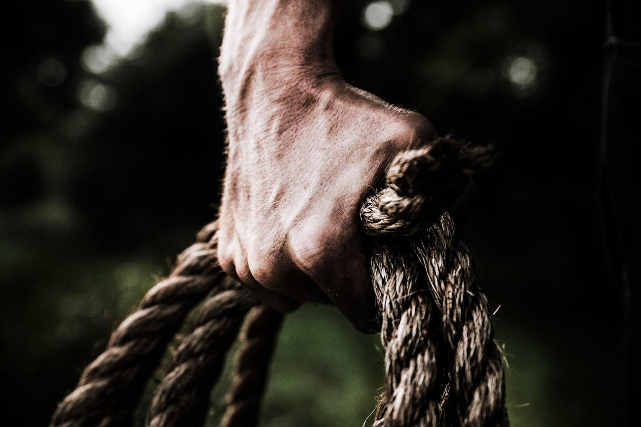 A close-up image shows a strong, firm hand gripping a thick, rough rope, showcasing strength and determination in an outdoor setting.
